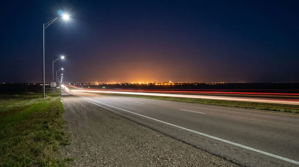 A four-lane highway at night with streaks of car lights and tall street lamps, the glow of a small city on the flat horizon.